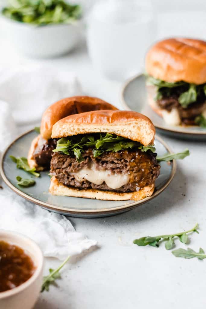 A lamb burger cut in half, with cheese oozing out, topped with fig preserves and arugula, with a brioche bun, on a blue plate