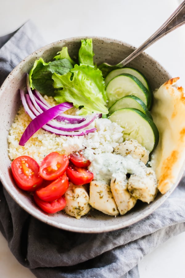 A chicken gyro bowl with chicken, couscous, fresh greens, tomatoes, feta, red onions, and cucumber.