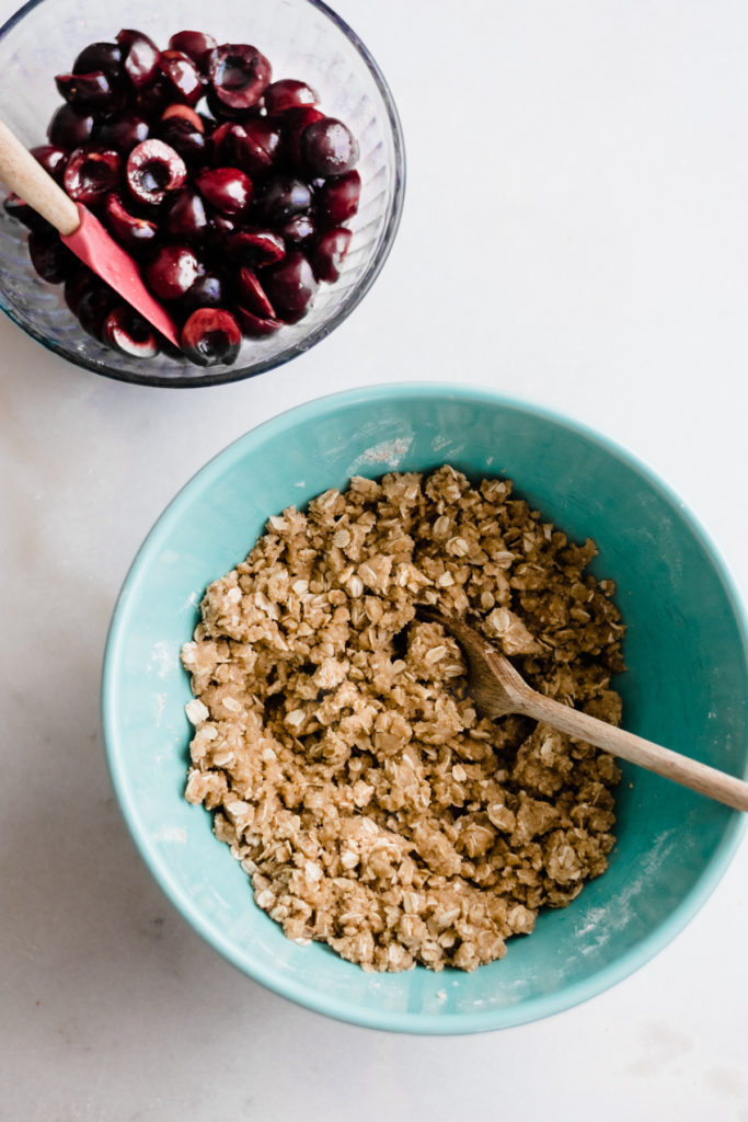 Halved cherries in a glass bowl and crumble bar mix in a blue bowl.