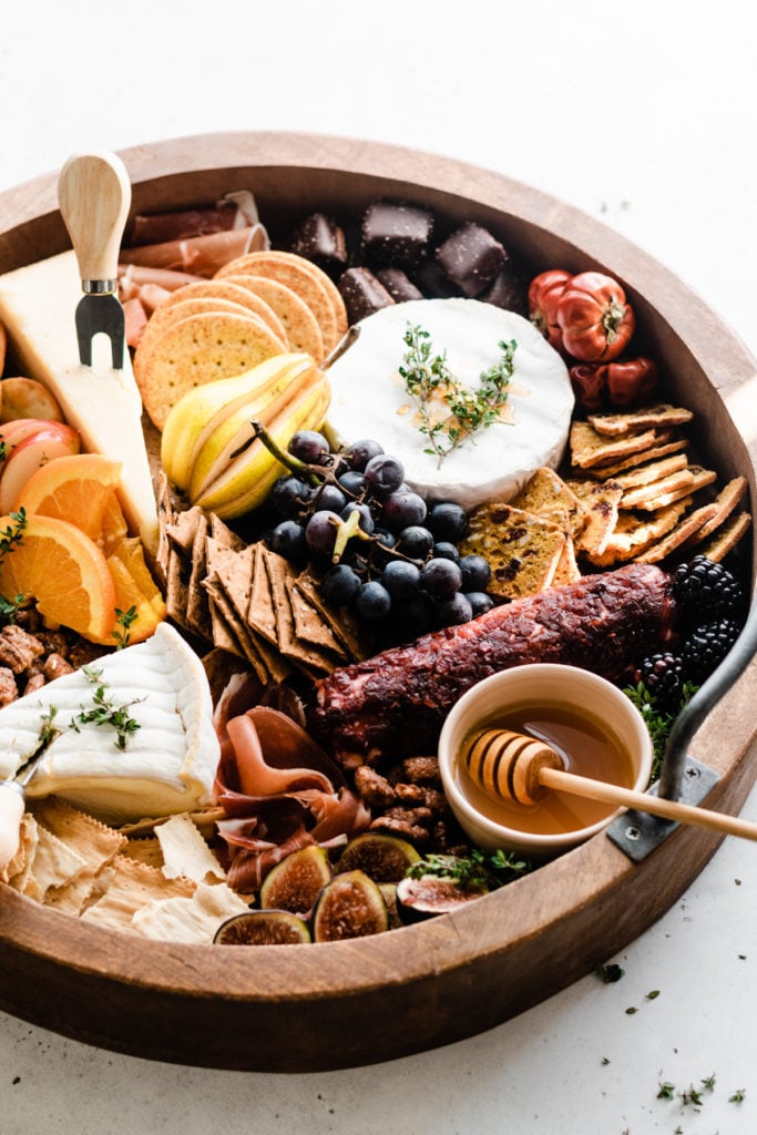 A round wooden board filled with fall produce, cheeses, crackers, meats, and jams.