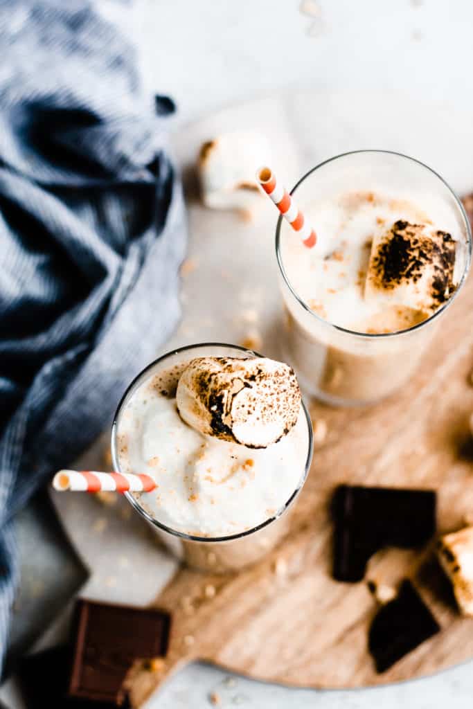 Bird's eye view of two tall glasses filled with toasted marshmallow milkshakes, on a wooden board, topped with toasted marshmallows and whipped cream and chocolate pieces laying nearby