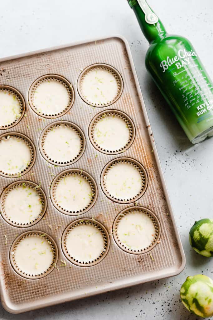 A muffin tin with muffin papers full of the unbaked cheesecakes, with zested limes and a bright green rum bottle on a neutral surface
