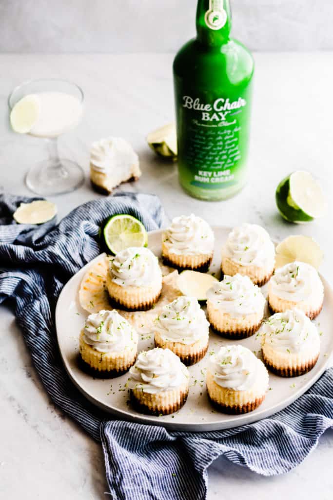 A plate of key lime cheesecakes on a blue linen, with a bright green Blue Chair Bay Key Lime Rum Cream bottle in the background. 