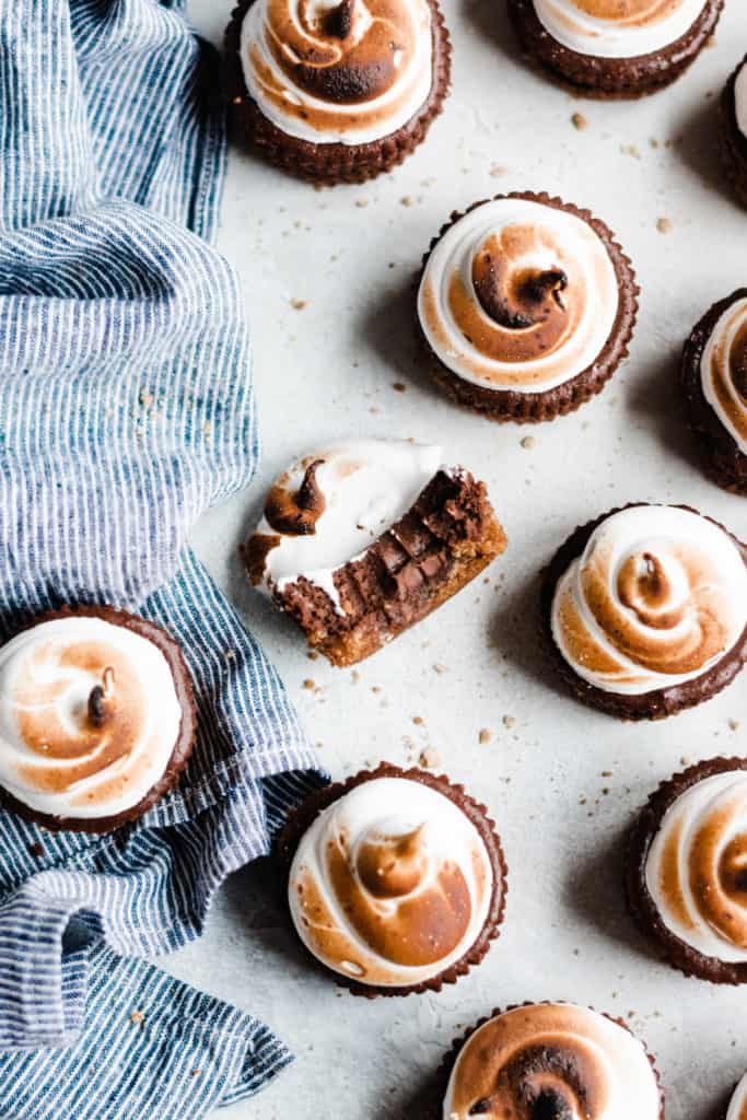 Mini Chocolate Cheesecakes topped with toasted meringue on a gray surface with a blue striped linen. One cheesecake has a bite taken out. 