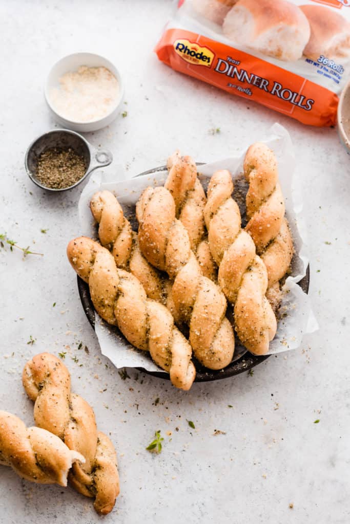 A plate of parmesan and herb sprinkled twisted breadsticks, with the Rhodes dinner roll bag in the background