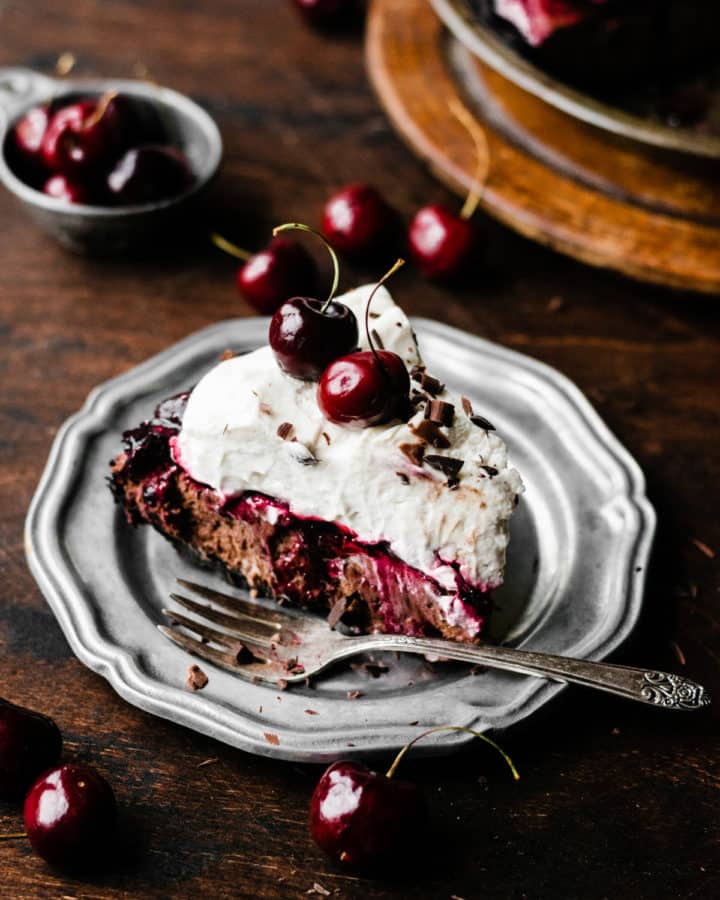 A slice of Black Forest Pie on a plate, with a fork and fresh cherries scattered around