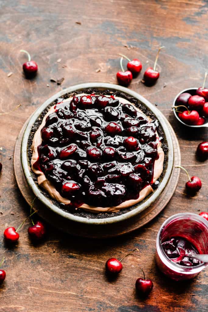 Chocolate mousse filling in a chocolate pie crust, topped with cherry sauce, on a wooden surface dotted with fresh cherries
