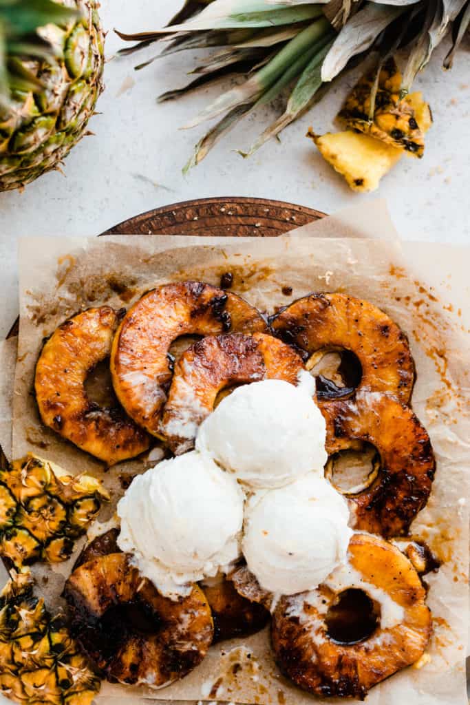 A close up of a wooden platter of grilled pineapple rings, topped with vanilla ice cream, with a pineapple laying nearby