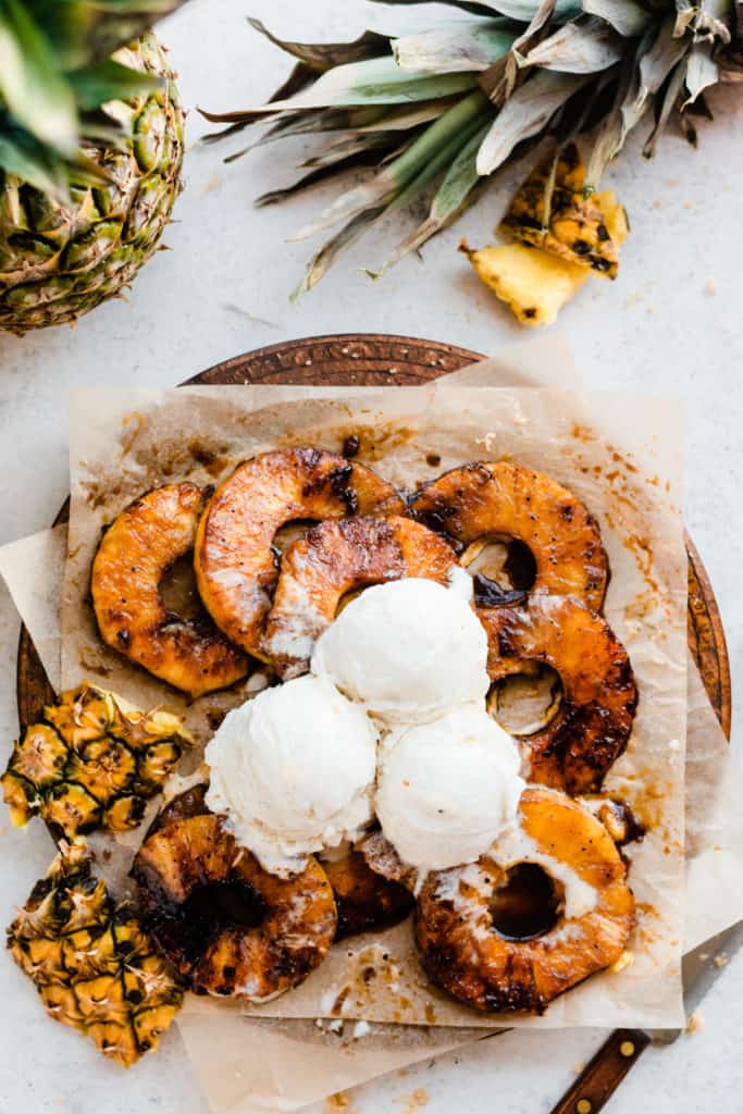 A wooden platter of grilled pineapple rings, topped with vanilla ice cream, with a pineapple laying nearby