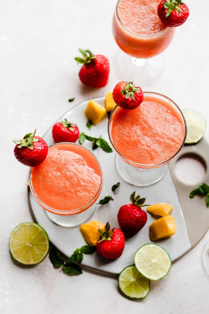 Three cocktail glasses from above, filled with a bright pinky orange punch and topped with fresh strawberries, with lime slices, mango chunks, and strawberries scattered around