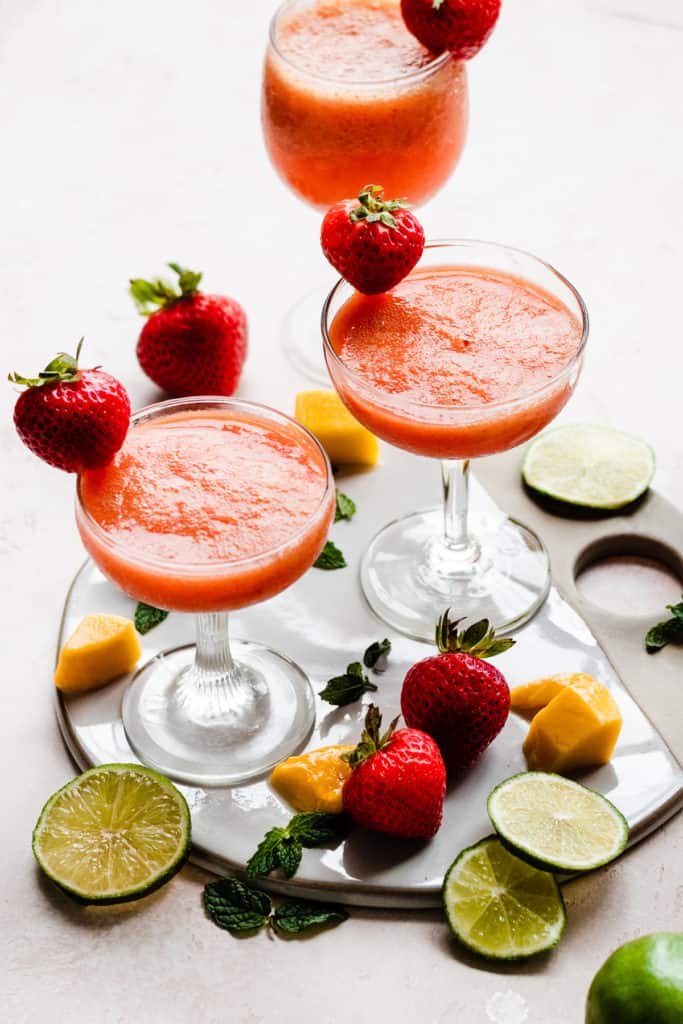Three cocktail glasses filled with a bright pinky orange rum punch, with mangos, strawberries, and lime slices scattered around