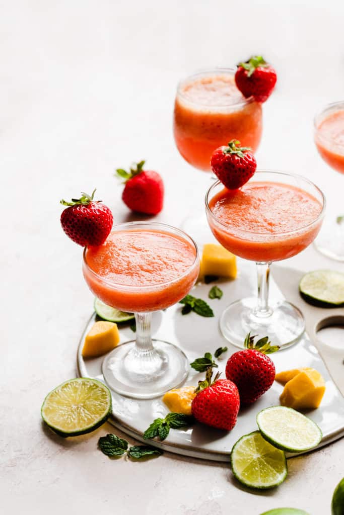 Four cocktail glasses filled with a bright pinky orange rum punch, with mangos, strawberries, and lime slices scattered around