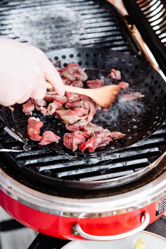 A hand holding a wooden spoon stirs beef strips in a grill pan on the grill