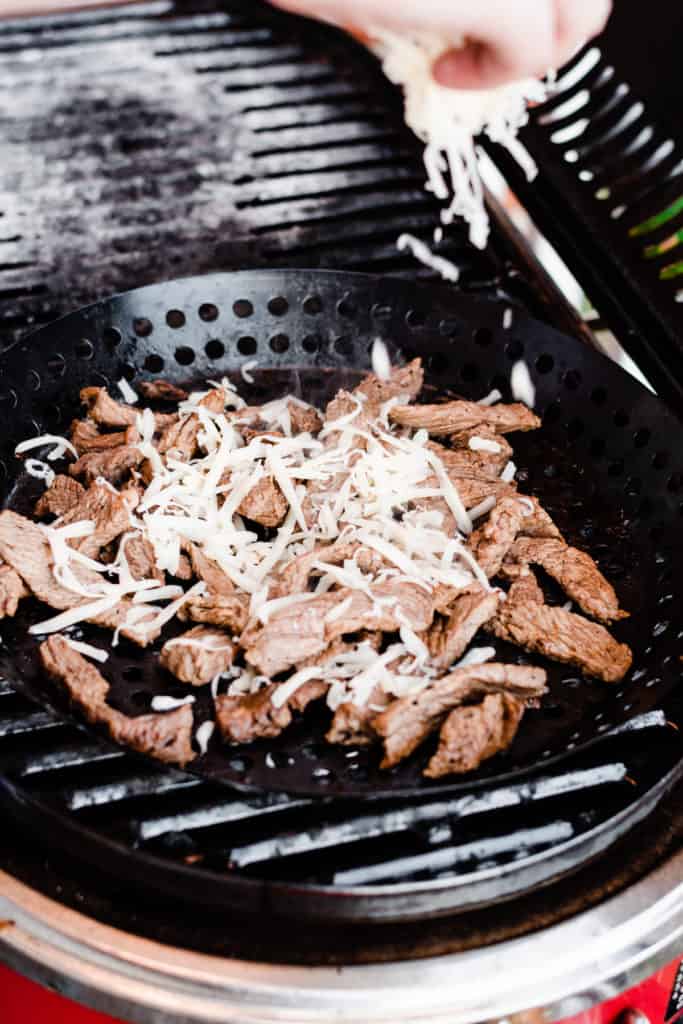 A hand sprinkles shredded Provolone cheese over the cooked beef strips in the grill pan