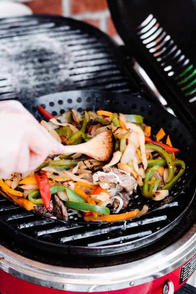 A hand stirs the veggie, onion, and beef mixture in the grill pan, with a wooden spoon