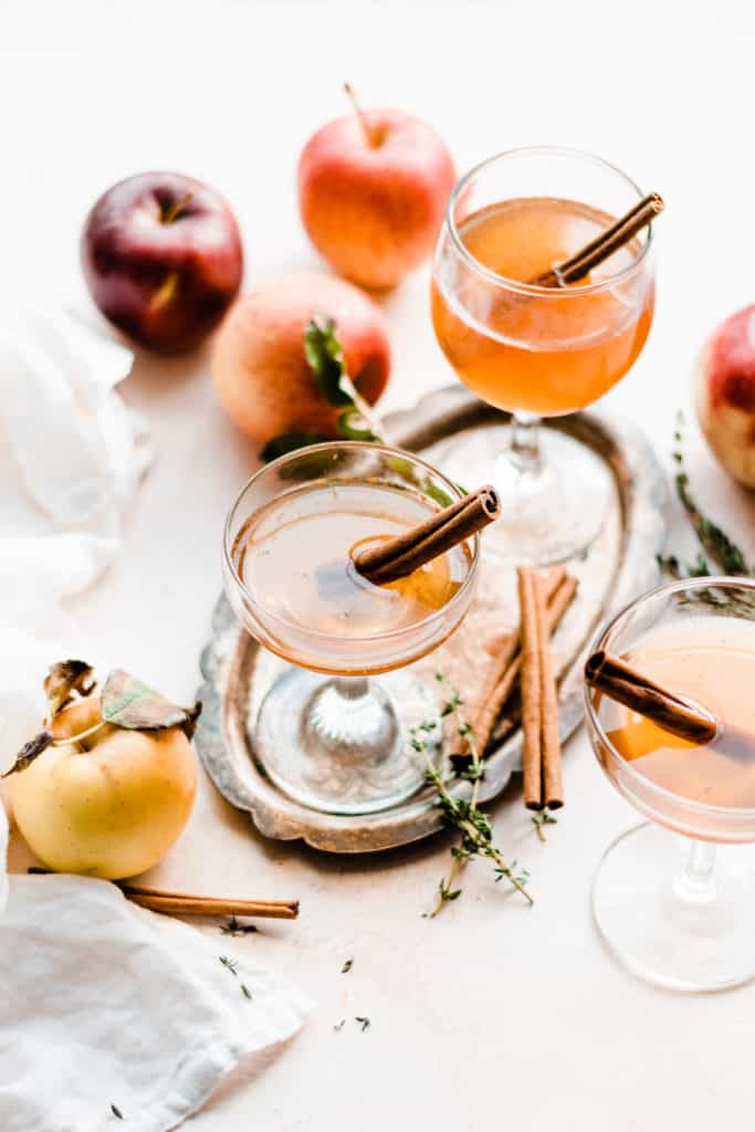 Three stemmed glasses filled with apple cider old fashioneds, with apples and cinnamon sticks scattered around