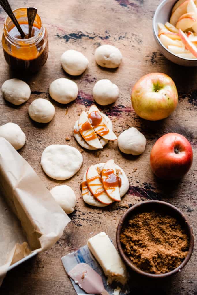 A wooden surface with dough balls, a jar of salted caramel sauce, apples, and apple slices. A few of the dough balls are flattened with brown sugar, apples, and caramel sauce on top. 