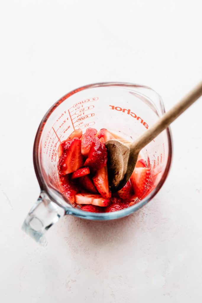 A glass measuring cup with sliced strawberries, with a wooden spoon muddling the berries