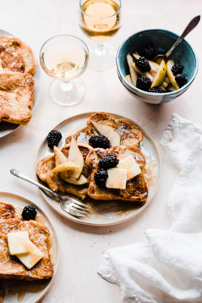 Two plates of golden french toast topped with butter, maple syrup, and blackberries and pear slices, on a light pink surface with two glasses of apple juice and a white linen. A bite has been taken from the french toast