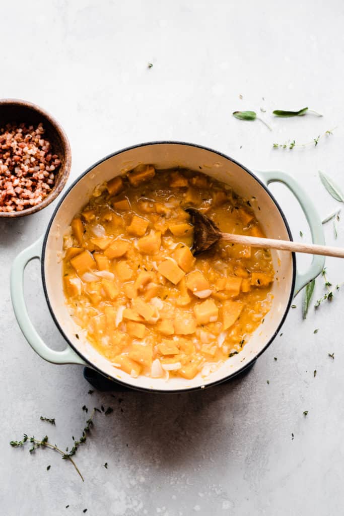 A dutch oven filled with cubed butternut squash and broth, with a bowl of pancetta nearby