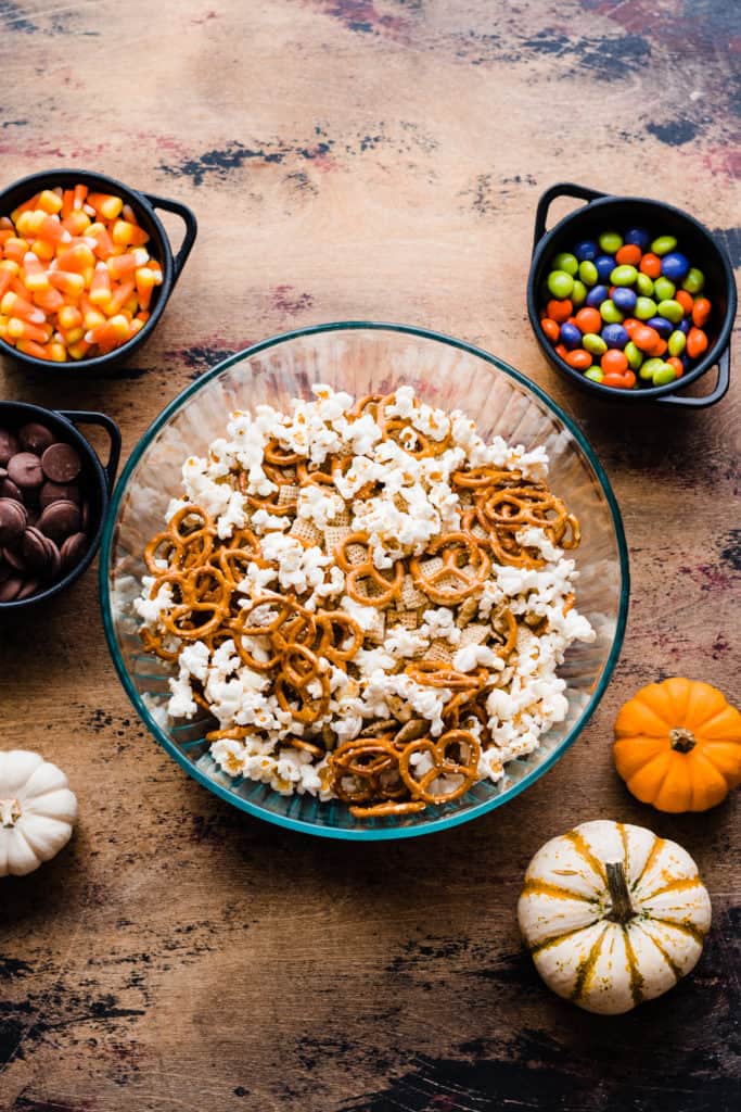 A bowl of popcorn, pretzels, and chex on a wooden surface surrounded by bowls of candy
