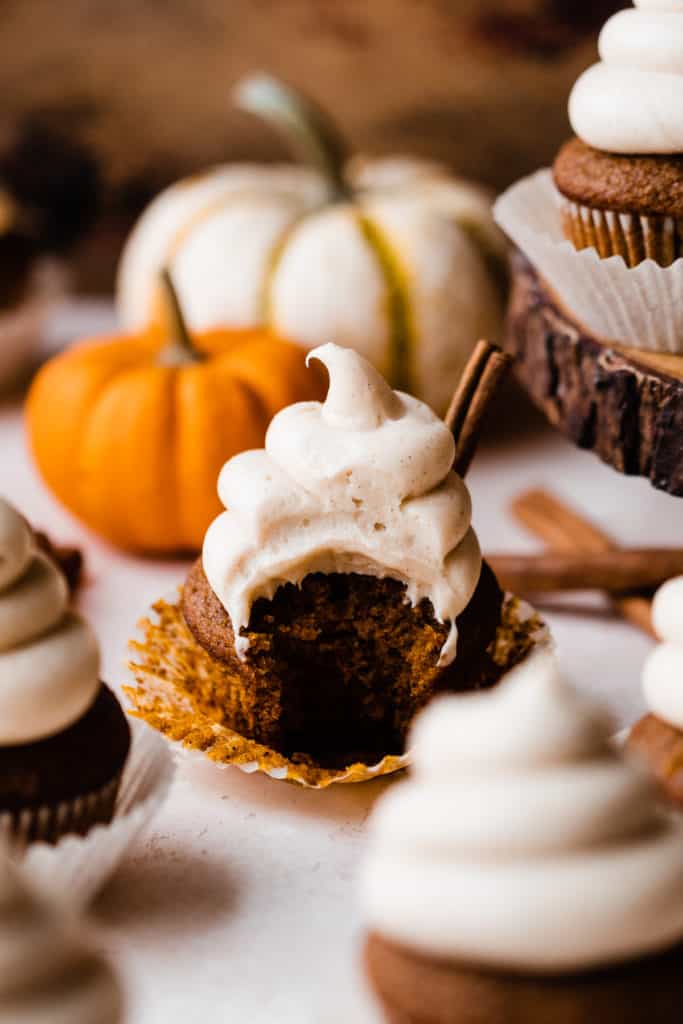 A pumpkin cupcake with a bite taken out, with mini pumpkins and cinnamon sticks in the background
