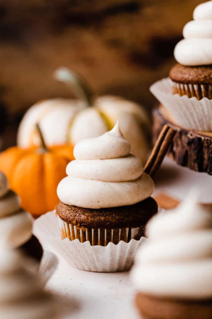 A close-up of a frosted pumpkin cupcake, with mini pumpkins behind it and a cinnamon stick in the cupcake