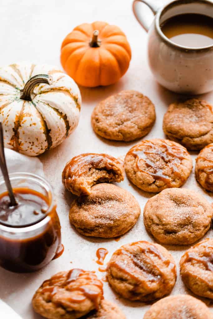 Pumpkin snickerdoodles on a light surface with mini pumpkins, a jar of caramel, and a mug of coffee