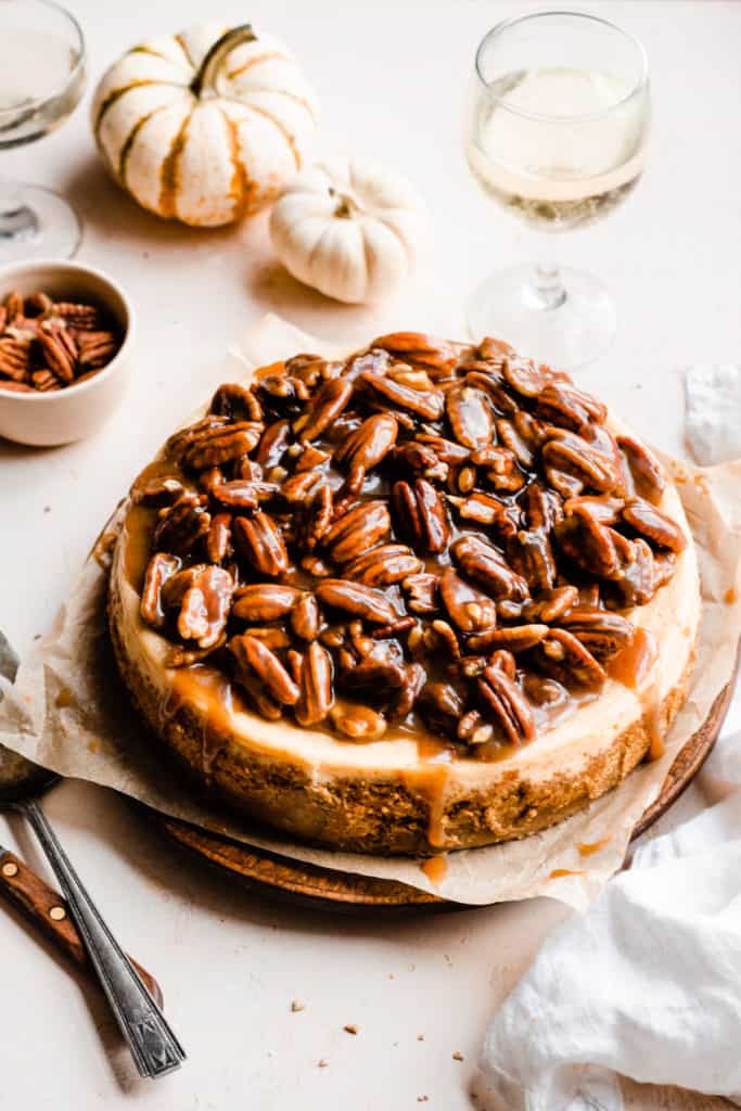 Pecan Pie Cheesecake with pecan topping and caramel drips, surrounded by mini pumpkins and wine glasses