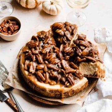Cheesecake topped with pecan praline, on a white background. A triangle of cheesecake has been sightly pulled back from the cake to show the interior of the sice.