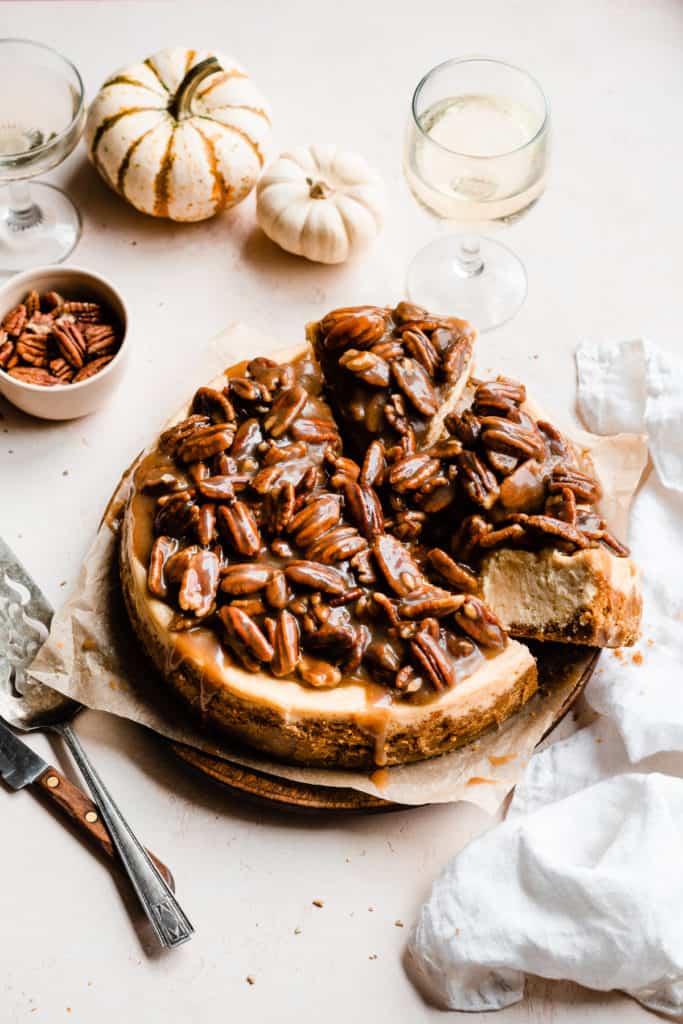 Pecan Pie Cheesecake with slices cut, surrounded by mini pumpkins, wine glasses, and a white linen