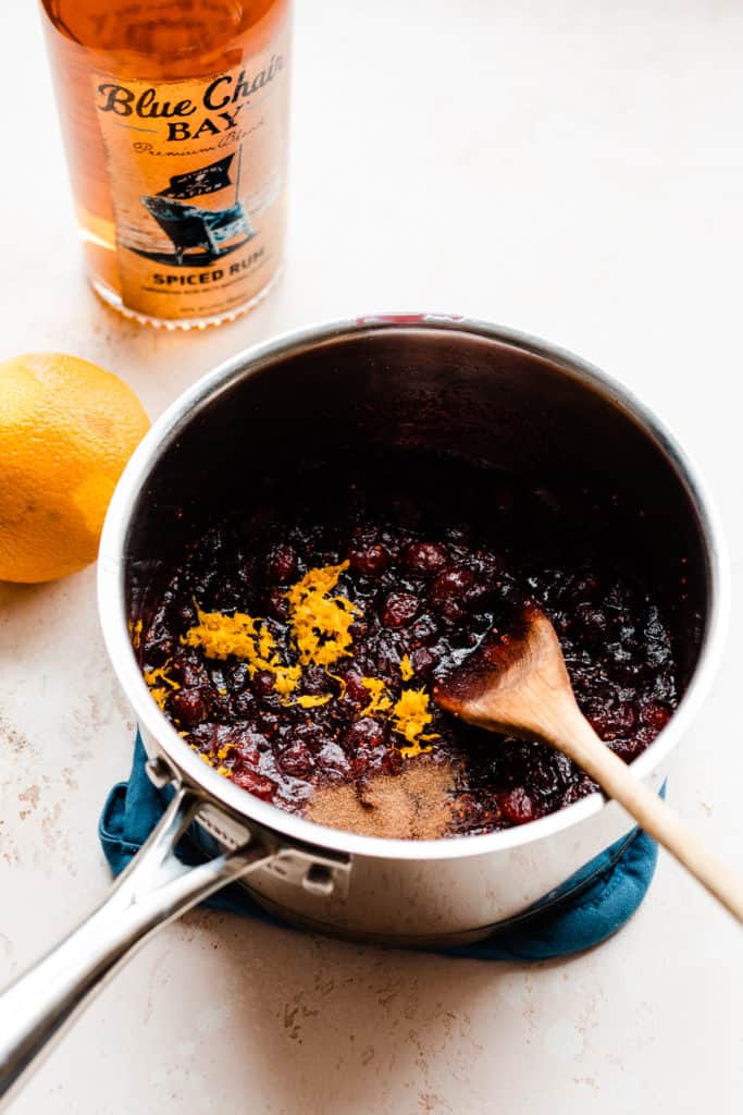 A pot of cranberry sauce with orange zest sprinkled on top, and a bottle of spiced rum