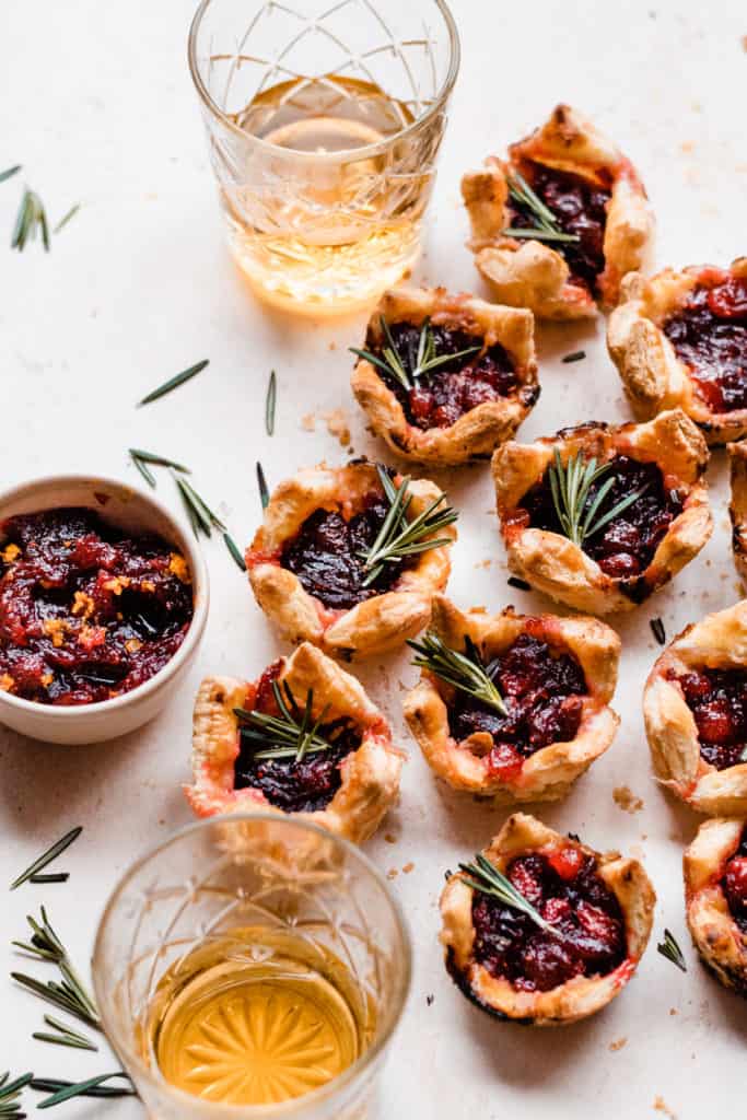 Puff pastry bites on a light surface surrounded by rosemary sprigs, glasses of rum, and a bowl of cranberry sauce