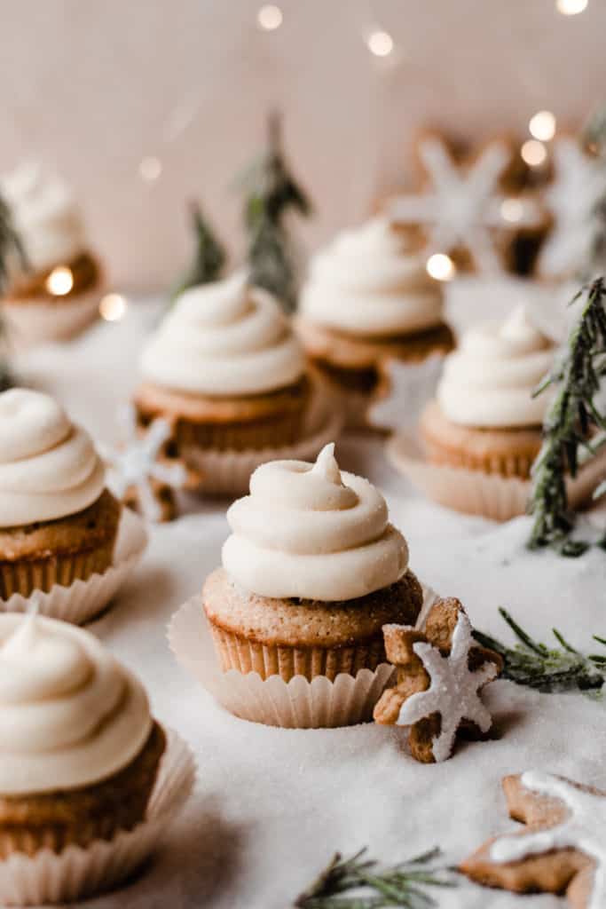 Cupcakes topped with swirls of cream cheese frosting on a bed of sugar "snow" with rosemary "trees and twinkle lights.