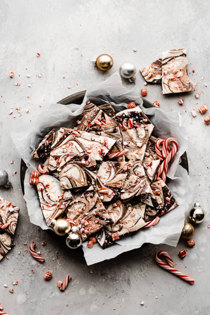A pie plate with parchment paper, filled with pieces of peppermint bark, with candy canes and mini ornaments scattered around