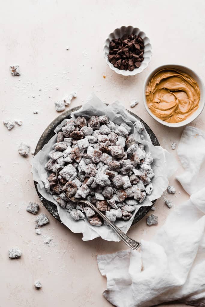 A dish of puppy chow, a bowl of chocolate chips, and a bowl of peanut butter on a light surface