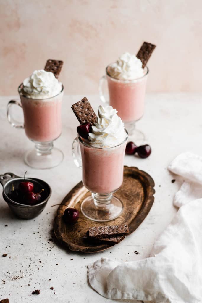 Three glasses of pink milkshake, topped with whipped cream, cherries, and chocolate graham crackers