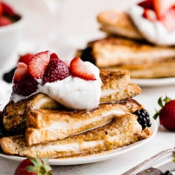 A close-up of a stack of french toast topped with whipped cream, berries, and maple syrup.