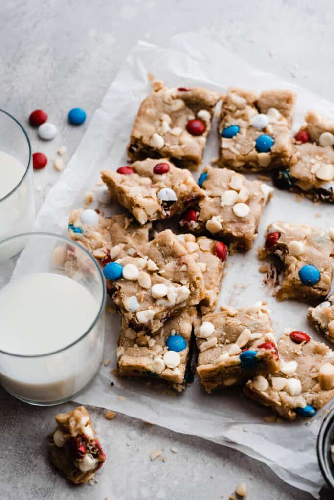 Stacked cookie bars on parchment paper with two glasses of milk nearby. 