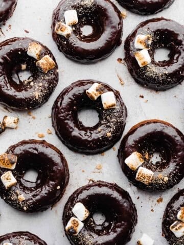 S'mores Donuts with chocolate ganache, toasted marshmallows, and graham cracker crumbs on a light gray surface.