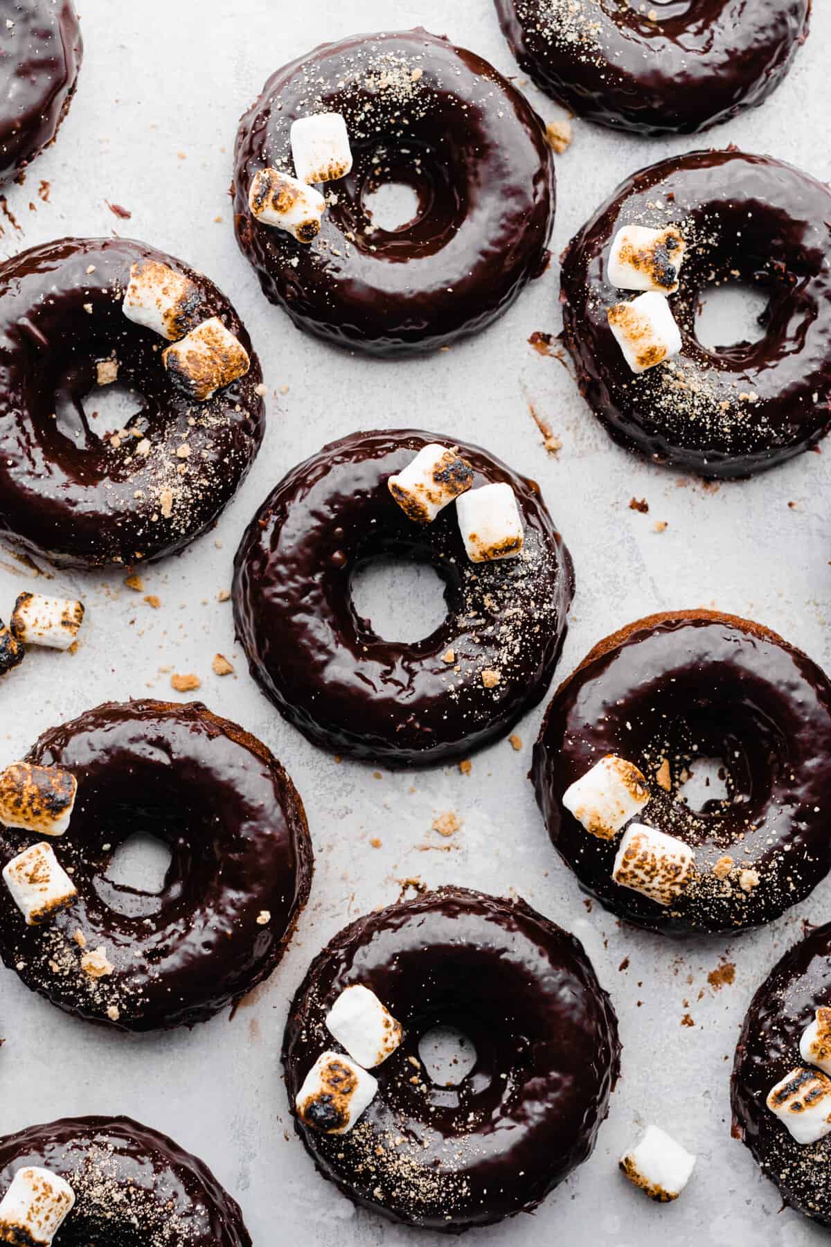 S'mores Donuts with chocolate ganache, toasted marshmallows, and graham cracker crumbs on a light gray surface.