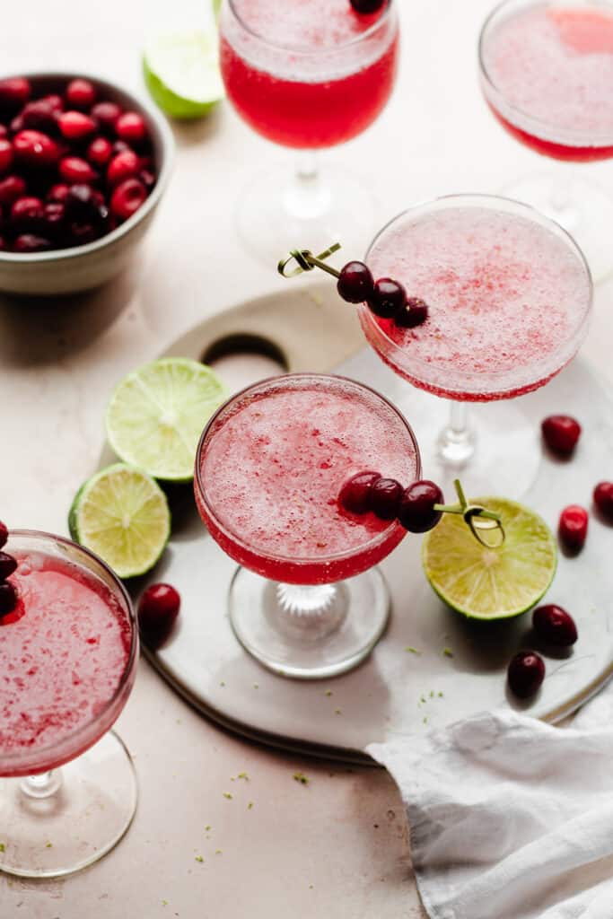 5 glasses of frozen cranberry daiquiris with cranberries and lime slices scattered around.