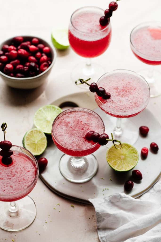 5 glasses of frozen cranberry daiquiris with cranberries and lime slices scattered around.