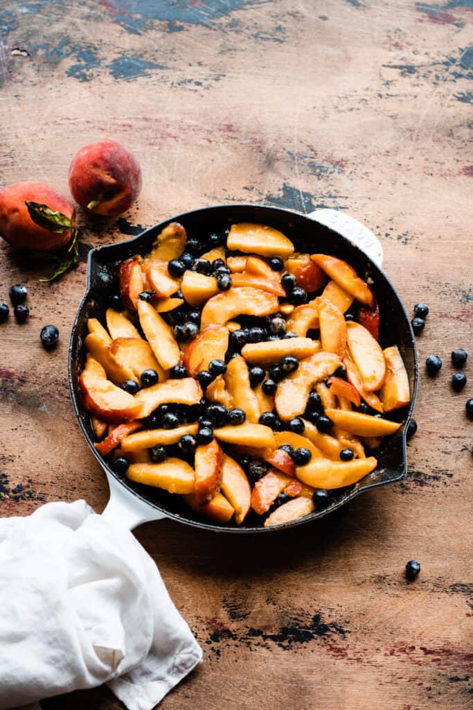 Blueberries and peach slices in a cast iron skillet on a wooden surface.