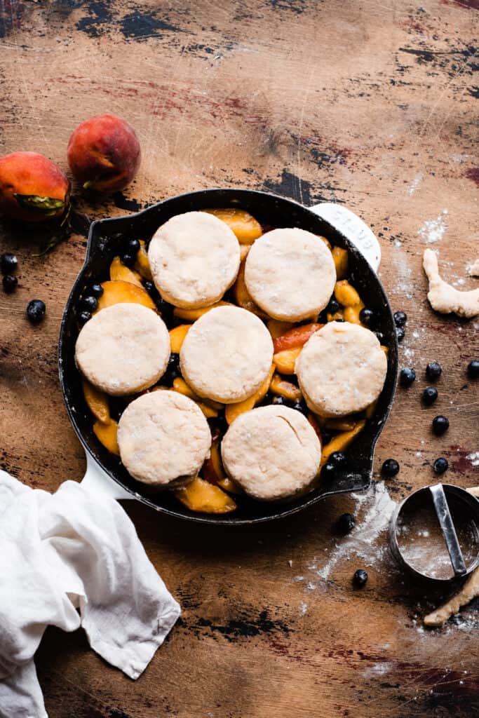 A cast iron skillet with blueberries and peach slices, topped with the unbaked biscuits.