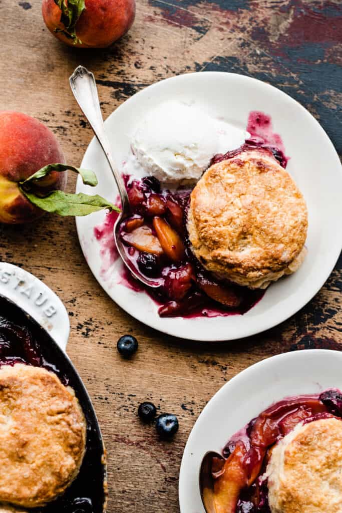 Two plates of the vibrant blueberry peach cobbler, with fresh blueberries and peaches scattered around, on a wooden surface.