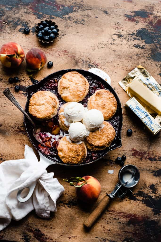 Blueberry Peach Cobbler in a cast iron skillet, topped with biscuits and vanilla ice cream, on a wooden surface.