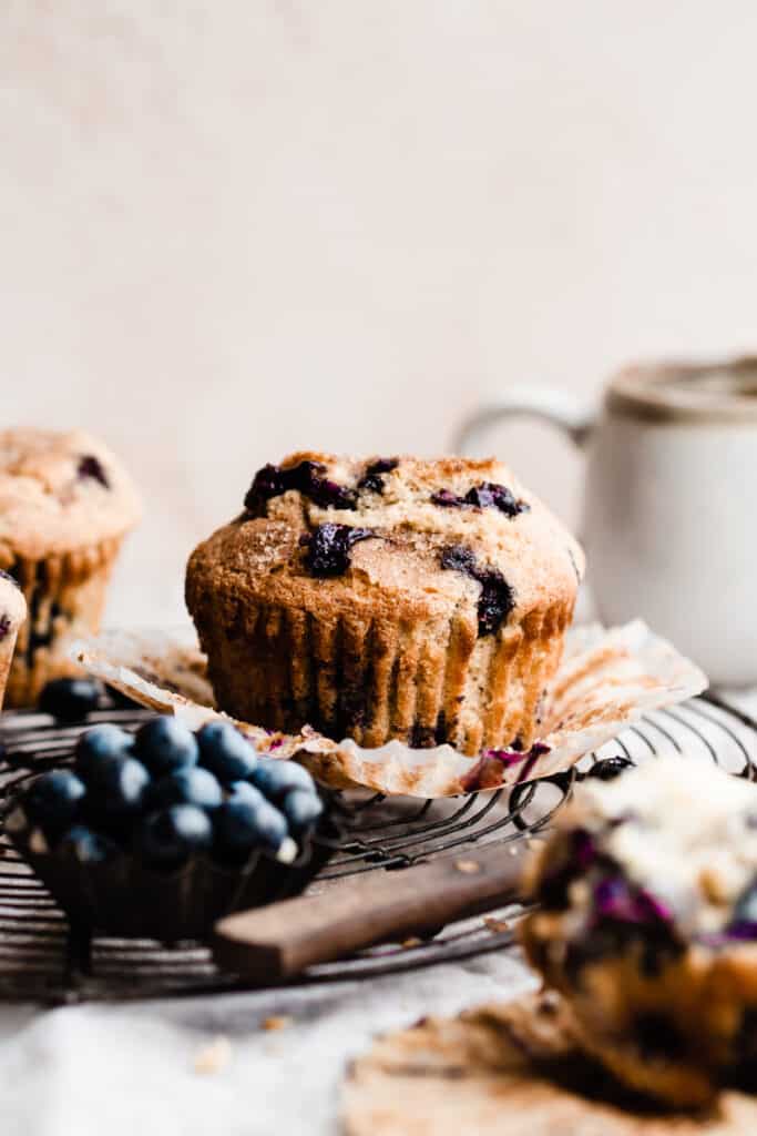 A blueberry muffin on a cooling rack with other muffins in the background, and a mug of coffee.