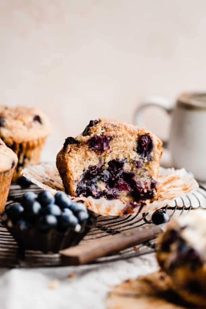 A blueberry muffin cut in half on a cooling rack, with blueberries and a coffee mug in the background.