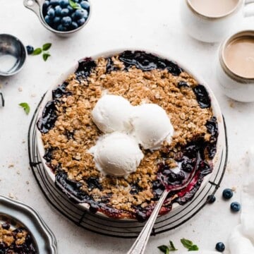 A round dish of blueberry crisp topped with ice cream, with a serving spoon digging in.